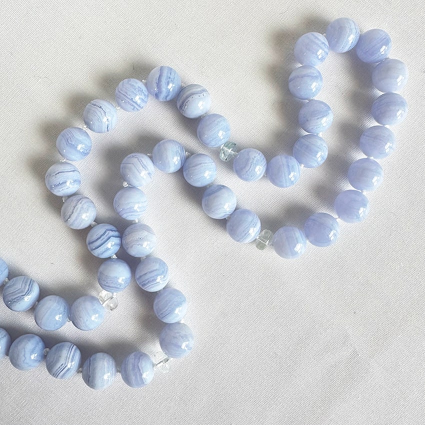 A close-up photograph of a blue lace agate bead necklace, featuring large, round beads with delicate white and lavender-blue bands, interspersed with small, clear faceted quartz crystal beads, laid upon a textured white linen background.