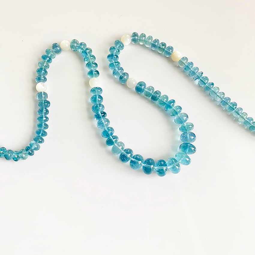 A long strand of aquamarine gemstone necklace featuring translucent, light-blue disk-shaped beads, interspersed with several contrasting opaque white moonstone beads, arranged in a curve on a plain white surface.