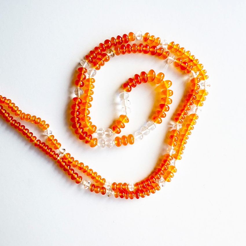An overhead view of a coiled necklace made of hundreds of small, vibrant orange-red carnelian disk-shaped beads, interspersed with small, clear quartz donut beads, laid on a clean white surface.