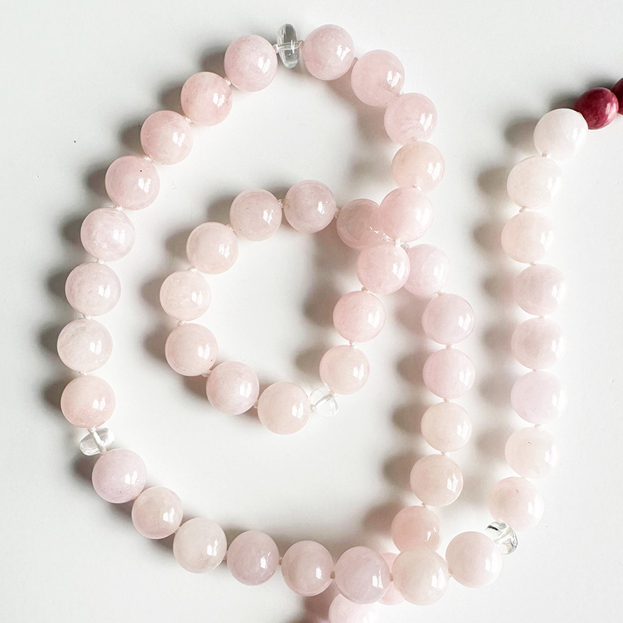 A coiled 10mm rose quartz bead necklace, featuring smooth, translucent soft-pink spherical beads with clear quartz accent beads, laid on a plain white surface.
