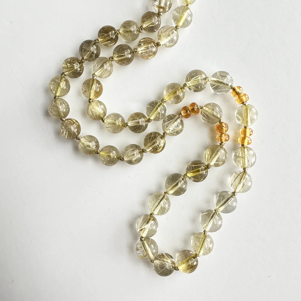 A close-up overhead view of a 6mm rutilated quartz bead necklace, showing round clear-to-smoky quartz beads with fine golden rutile inclusions, accented by small sections of golden-orange spessartite garnet beads, laid on a white surface.